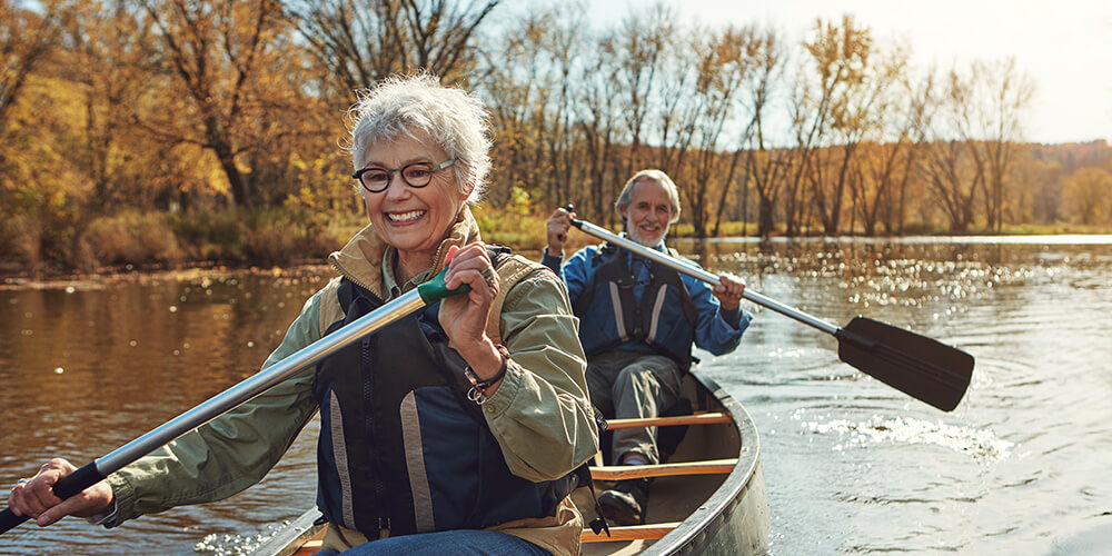 Two older adults paddle a canoe on a calm river surrounded by autumn trees. Both are smiling and wearing life jackets, enjoying the sunny fall day.