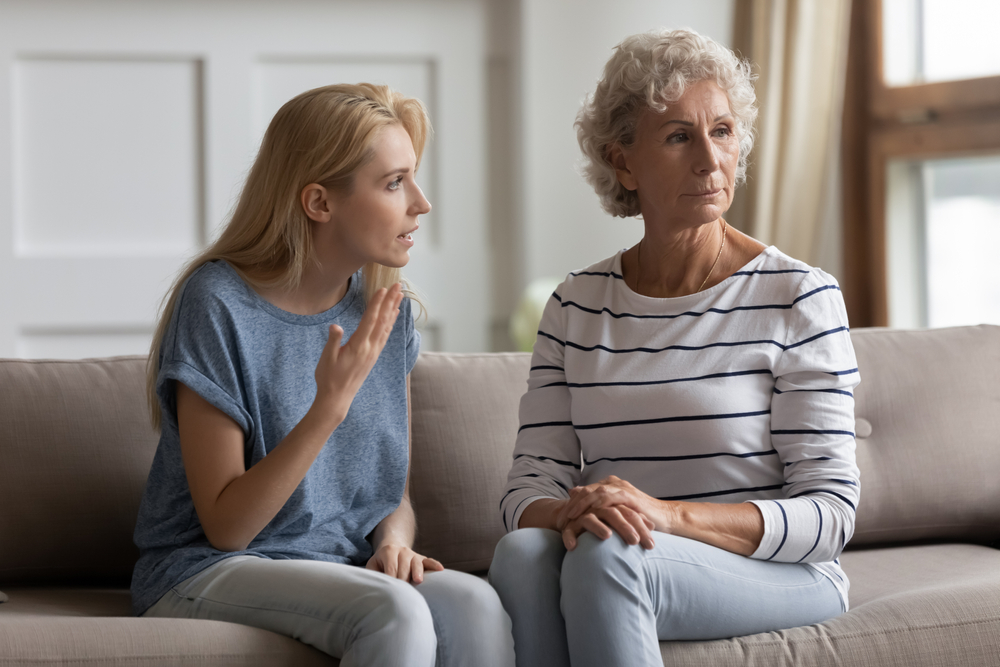 A younger woman talks to an older woman on a couch. The older woman looks away with a serious expression, while the younger woman gestures as she speaks, appearing frustrated or upset.