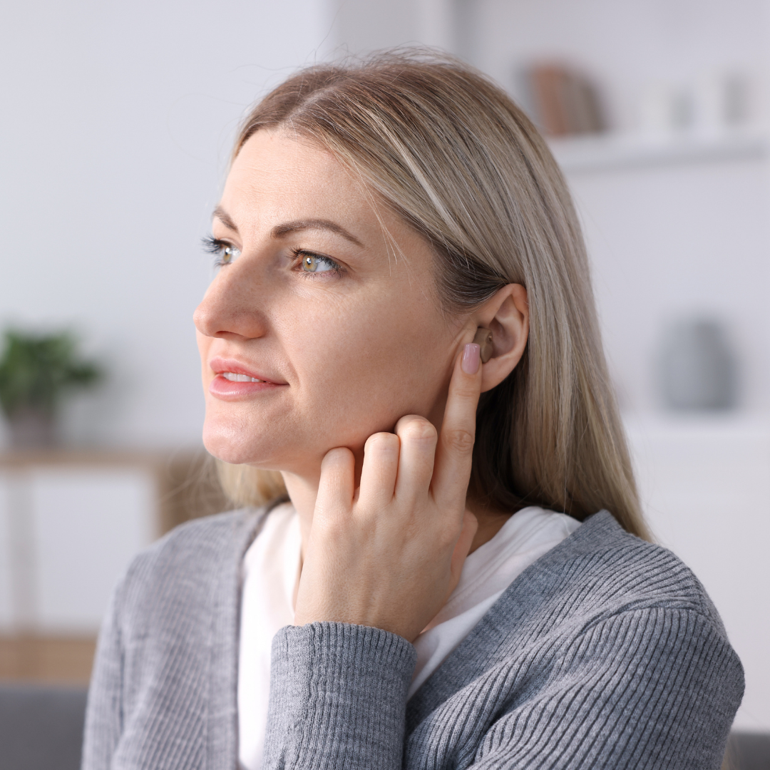 A woman with blonde hair wearing a gray sweater touches her ear with one finger while looking to the side, sitting indoors in a softly lit room.